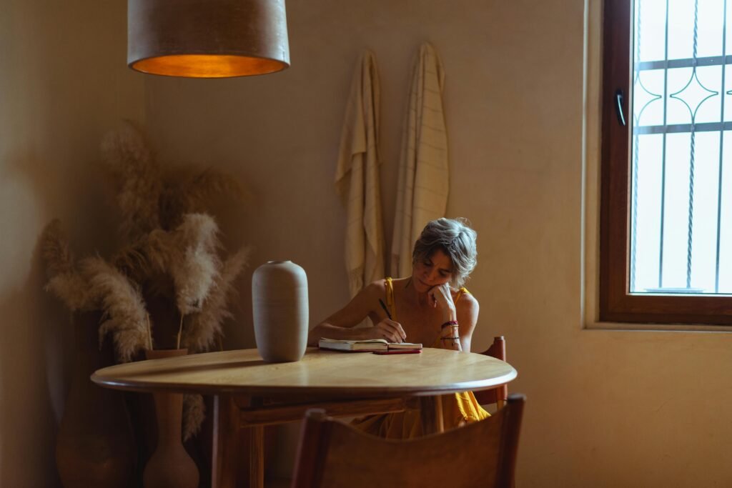 A senior woman enjoys journaling at a table in a warm, cozy home setting.